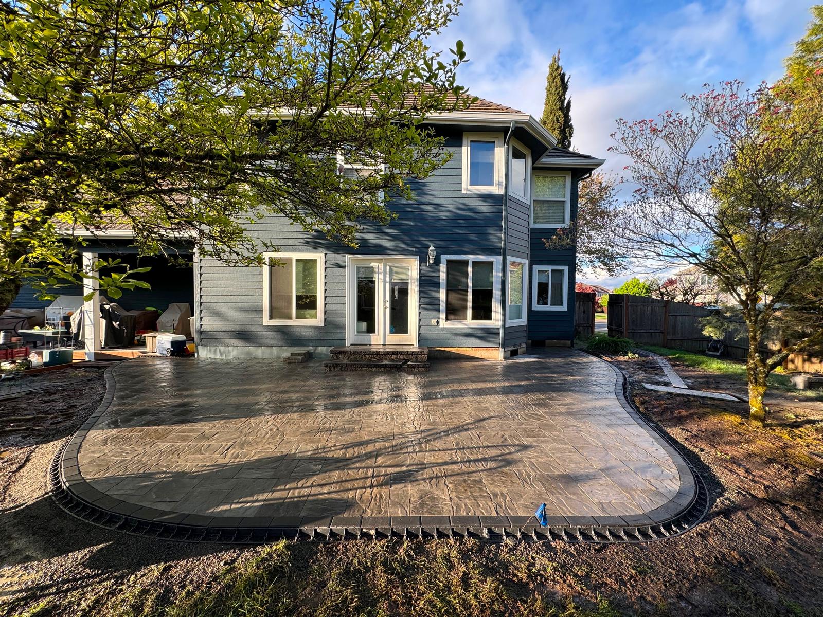 Finished stamped concrete patio with a dark charcoal border curving around the back of a blue two-story home